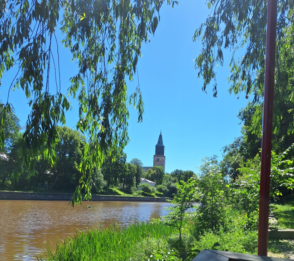 Joki kulkee keskellä kuvaa. Taustalla näkyy kirkon torni. Ympärillä ja yläpuolella vihreitä kasveja.
